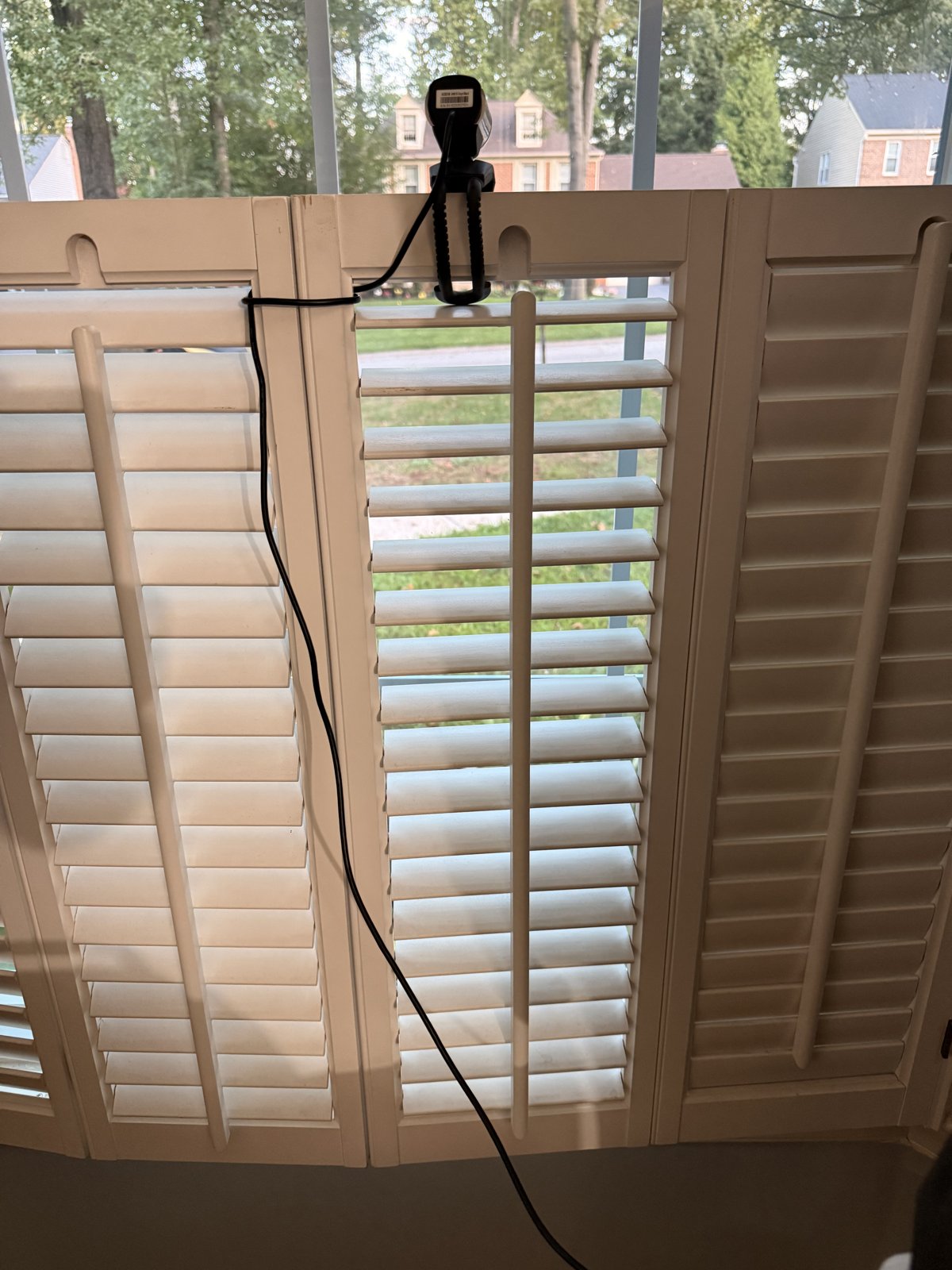 A small black USB webcam clamped to the top of white plantation shutters, pointed out a window at a tree-lined suburban street with houses across the way